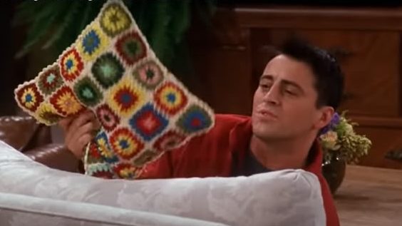 Ross holds up a crochet pillow covered in small, multi-colored granny squares.