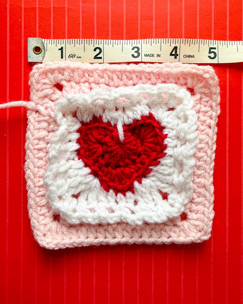 A red, pink, and whie granny square with a heart in the center and white lace details is shown on a red and light pink striped background. A measuring tape shows the square measures 5 inches wide.